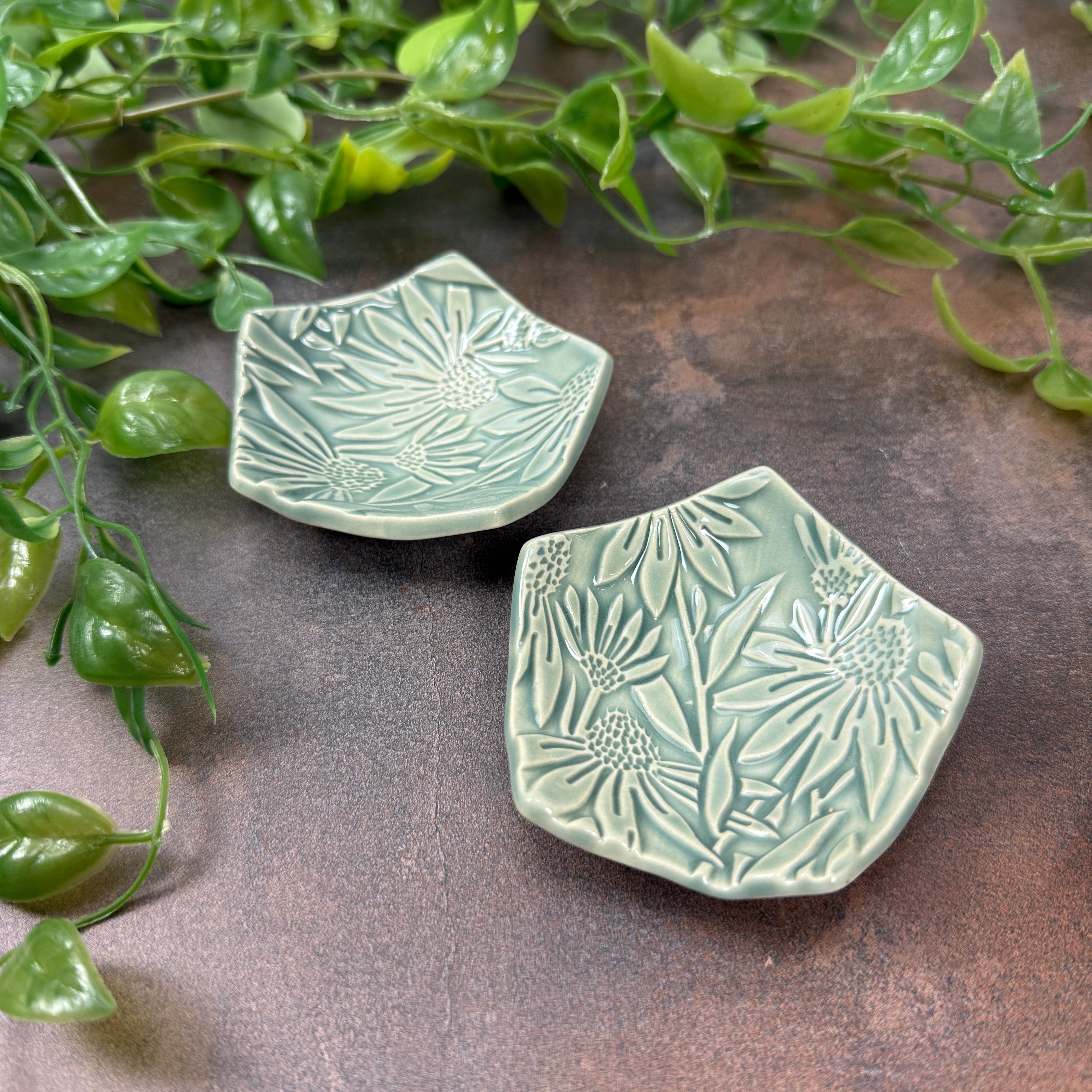 Two fog blue ceramic tea bag holders with leaf and flower patterns on a textured brown surface, surrounded by green leaves.
