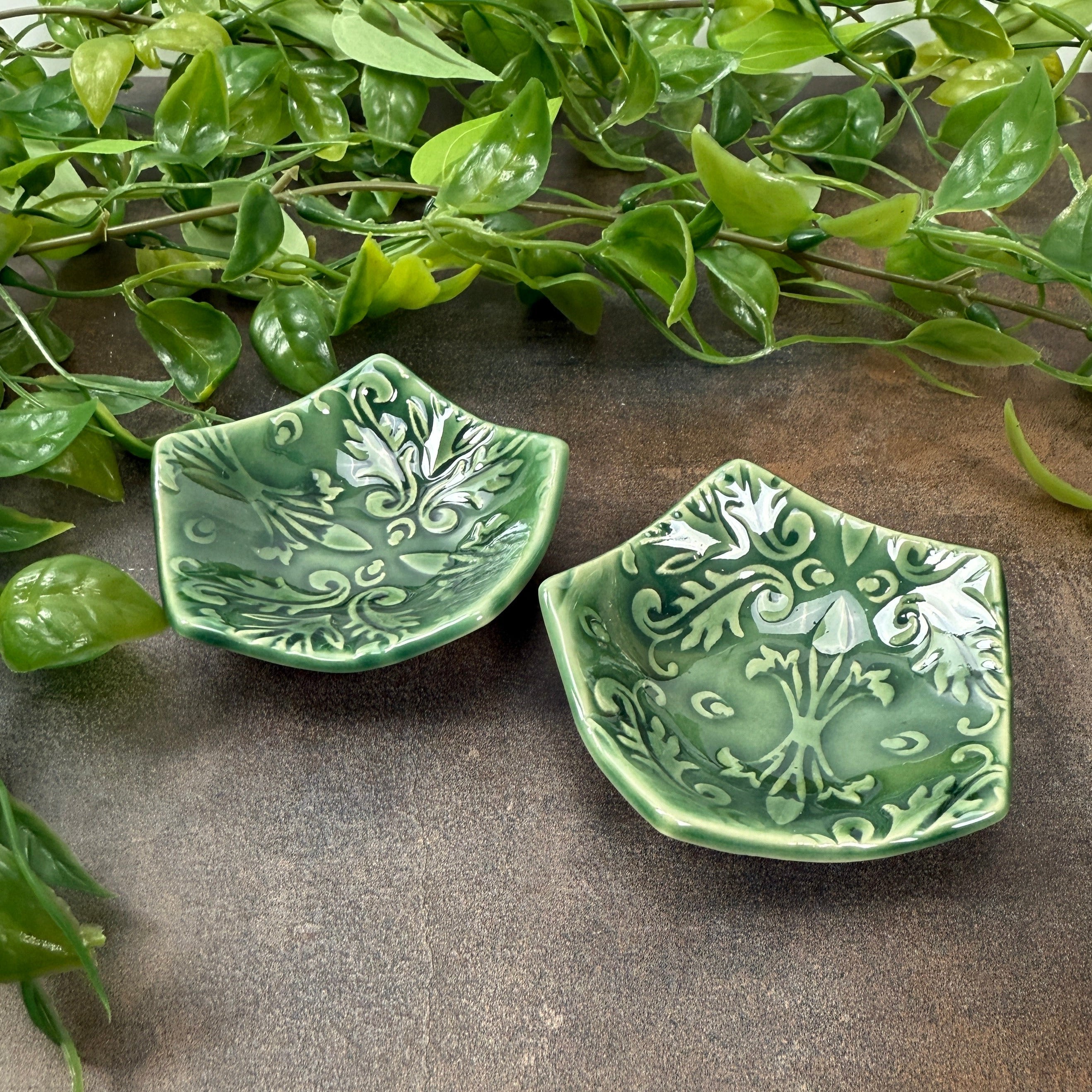 Two green ceramic tea bag holders with intricate patterns on a textured brown background.